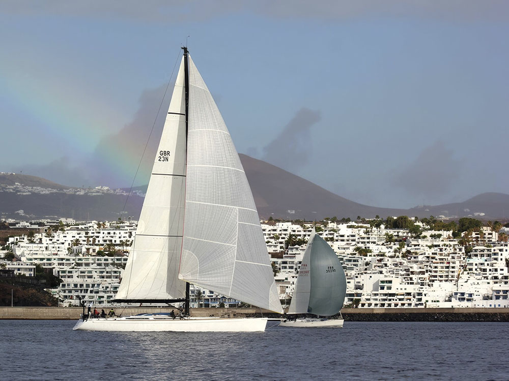 A sailboat on the water. In the background is a townscape packed with small white buildings and a rainbow in the sky