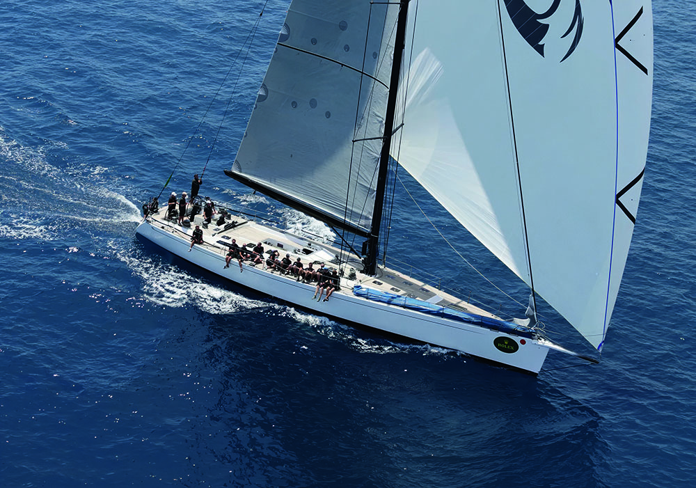 A sailboat on the water as taken from above. Many of the crew are sitting on the side with their legs dangling over the water