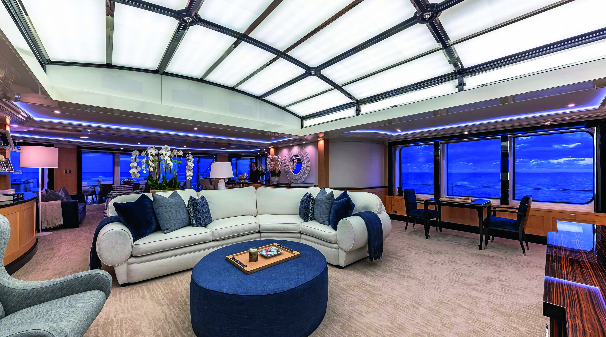 Lounge area with neutral curved sofa with blue cushions and blue table in the centre. The windows are tinted royal blue