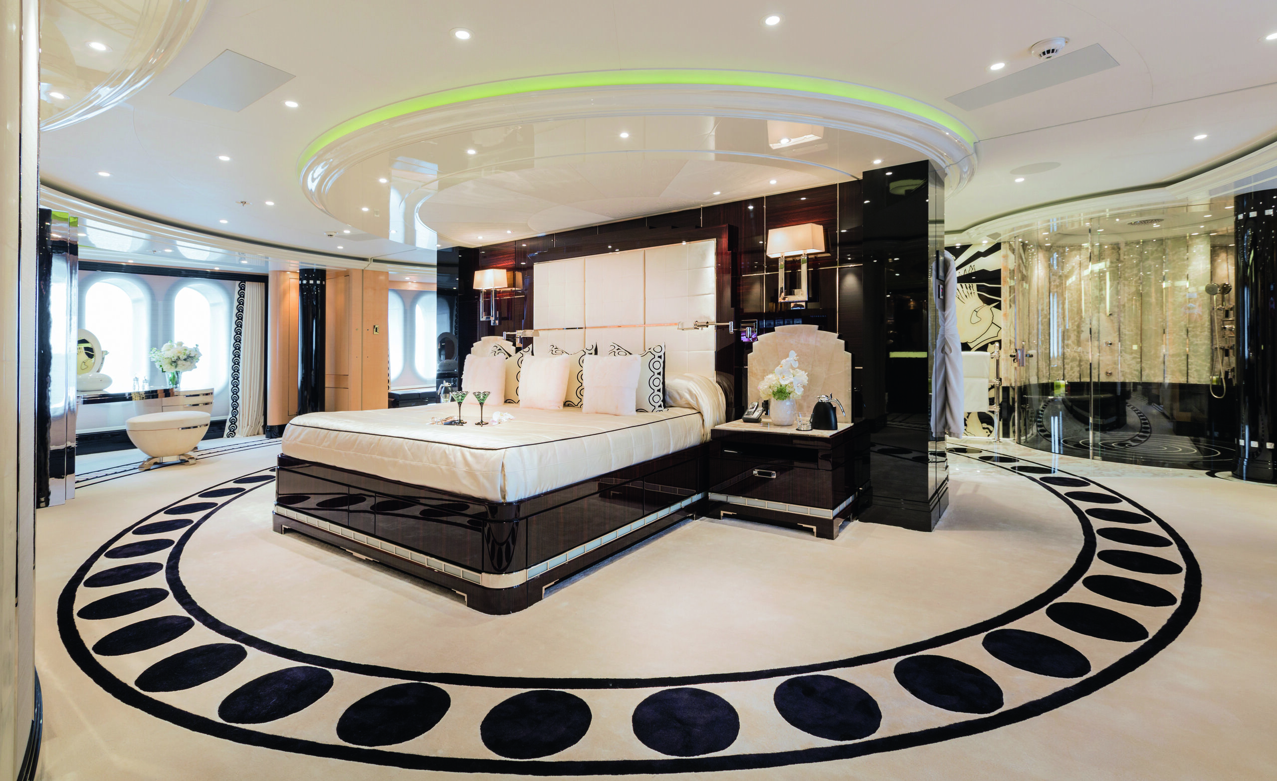 A large, black circular pattern on white-carpeted floor. At the centre is a bed with shiny black surround. Above is green lighting and behind is a large, open glass-fronted shower
