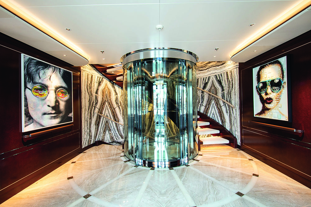 Glass-sided circular lift at the centre of a hallway with white marble flooring and a curved staircase behind